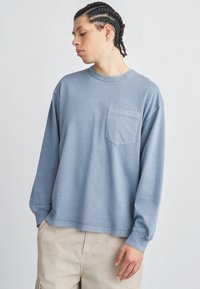 Ανοιχτό μπλε μακρυμάνικη μπλούζα από απαλό ύφασμα· διαθέτει στρογγυλεμένο λαιμό και μπροστινή τσέπη. Φορεμένη με μπεζ παντελόνι cargo.