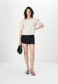 Lys beige bluse med blomsterdesign og pufferm, kombinert med et svart, folderet miniskjørt. Modellen holder en svart håndveske og har på seg sandaler med remmer.