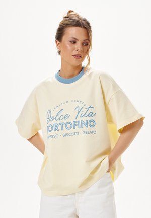 Młoda kobieta z włosami związanymi w kok, ubrana w kremowy, oversize T-shirt z niebieskim napisem oraz białe spodnie, stojąca z rękami w kieszeniach na białym tle.