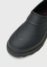 Black slip-on shoe with a smooth, matte finish. Rounded toe and thick, textured sole for grip. Minimalist design with no visible hardware.