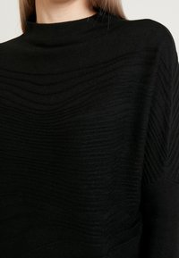 Svart höghalset tröja med strukturerade vågmönster, tillverkad av mjukt material, med en avslappnad silhuett och sänkta axlar.