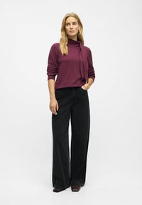 Bordeaux striktrøje med en knyttet halsudskæring, parret med løse sorte jeans og brune hæle. Model står med hænderne i lommerne.