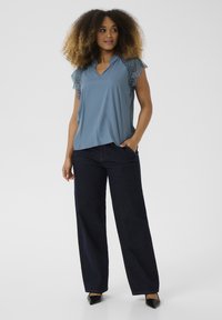 Blusa blu con maniche in pizzo, abbinata a pantaloni a gamba larga in denim scuro a vita alta e scarpe nere a punta.