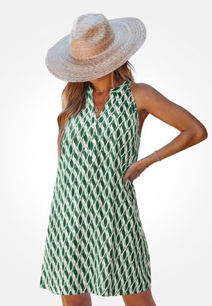 Mouwloze groene en witte jurk met patroon, gedragen met een strohoed met brede rand, gouden kettingen en armbanden, hand rustend op de heup.