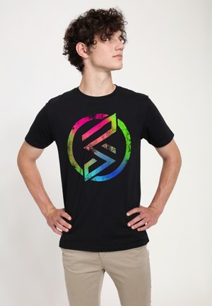 Sort bomulds T-shirt med et stort, flerfarvet cirkulært logo med en slidt tekstur, båret med lyse bukser.
