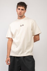 Camiseta de manga corta color crema de algodón con cuello redondo, con letras oscuras en el centro. Llevado con pantalones sueltos negros.