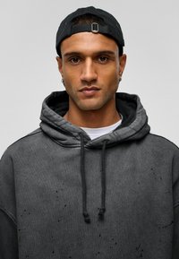 Donkergrijze hoodie met een gestructureerd oppervlak, voorzien van een kangoeroezak, trekkoorden en lichte slijtage. Gedragen met een zwarte pet.