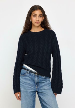 Kvinde med langt krøllet hår iført en mørk kabelstrikket sweater stukket ned i lyseblå jeans med et sort fletbælte mod en ensfarvet baggrund.