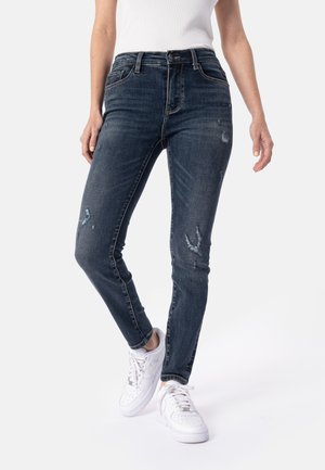 Person trägt dunkelblaue Skinny-Jeans mit leichter Abnutzung und weiße Sneaker, steht vor einem schlichten Hintergrund.