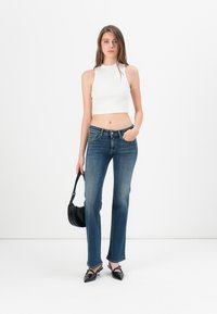 Sieviete baltā bezpiedurkņu topā, zilos džinsos un melnās norādītās kurpēs, turot nelielu melnu somiņu uz baltas fona.