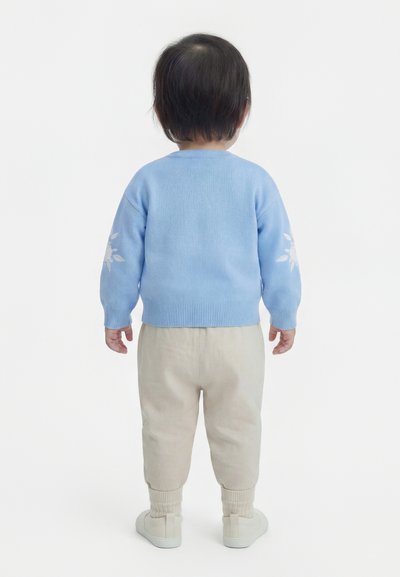 Νήπιο που κοιτάζει προς τα πίσω, φοράει ανοιχτό μπλε πουλόβερ με λευκά σχέδια φύλλων στα μανίκια, μπεζ παντελόνι και λευκά παπούτσια σε λευκό φόντο.