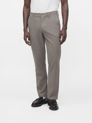 Homme portant un pantalon droit marron clair, des chaussettes blanches, des chaussures de ville noires brillantes et une chemise blanche sur un fond uni.