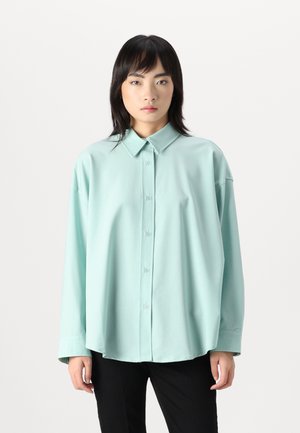 Lys teal oversized skjorte laget av et glatt stoff, med knappestolpe foran, klassisk krage og lange ermer. Kombinert med svarte bukser.