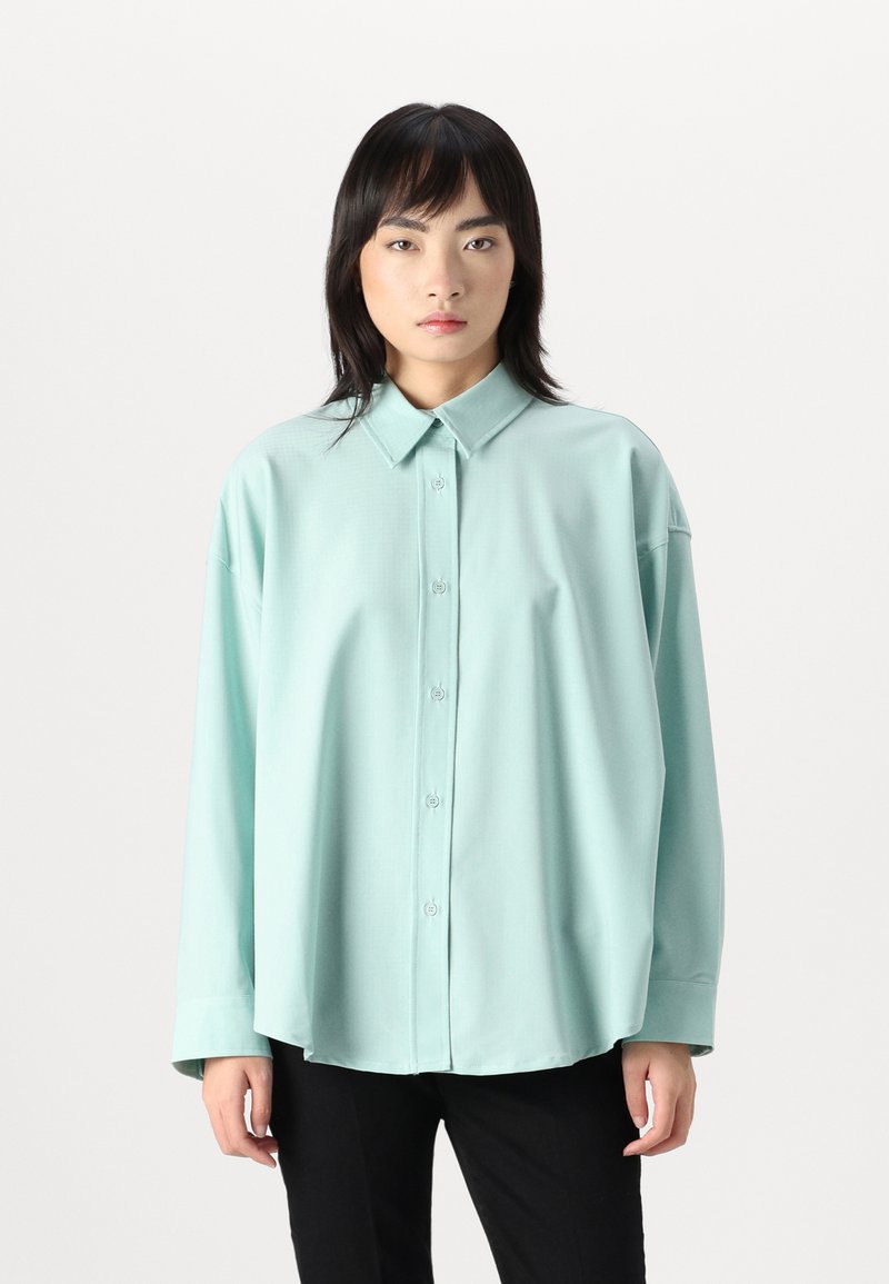 Camicia oversize di un blu acqua chiaro realizzata in un tessuto morbido, con chiusura a bottoni, colletto classico e maniche lunghe. Abbinata a pantaloni neri.