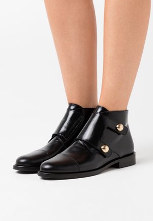 Botas negras de cuero brillantes hasta el tobillo con puntas afiladas, correas anchas y dos grandes botones dorados a cada lado, lucidas sobre piernas desnudas.