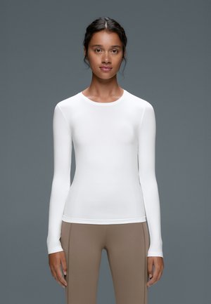 Femme aux cheveux foncés attachés, portant une chemise blanche cintrée à manches longues et un pantalon beige, debout devant un fond gris.