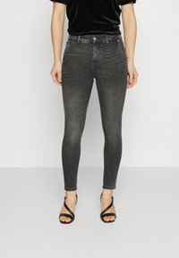 Mujer vestida con jeans ajustados negros, top de terciopelo negro y sandalias de tacón alto negras con tiras, de pie frente a un fondo blanco liso.