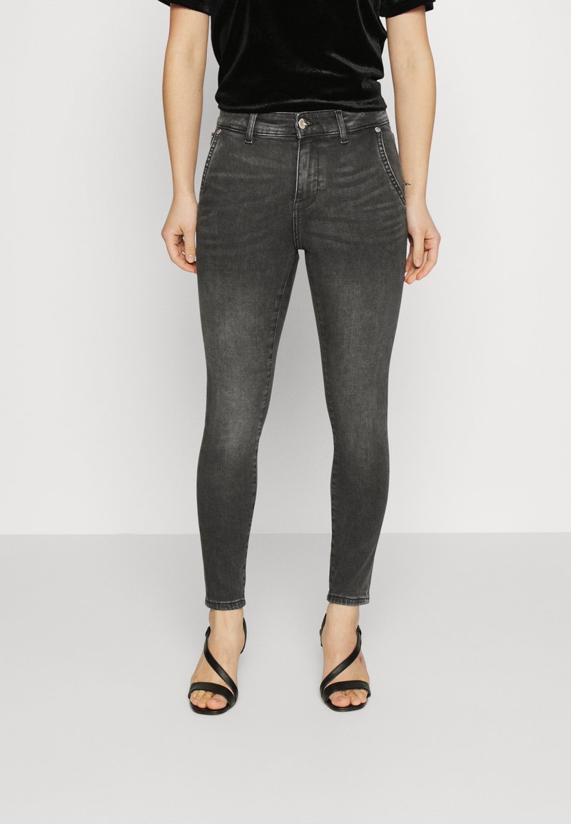 Mujer vestida con jeans ajustados negros, top de terciopelo negro y sandalias de tacón alto negras con tiras, de pie frente a un fondo blanco liso.