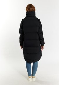 Schwarze Daunenjacke mit hohem Kragen, ausgestattet mit einem gesteppten Design und langer Silhouette. Getragen über blauen Jeans und weißen Sneakern.