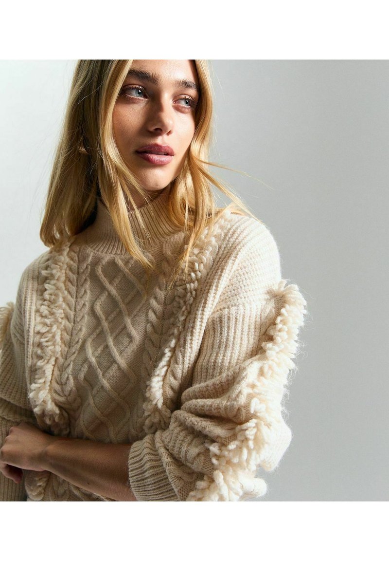 Maglione beige lavorato a maglia con motivi a treccia, dettagli testurizzati, colletto alto e frange sulle maniche. Fibra morbida e accogliente.