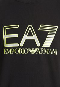 Musta t-paita, jossa on suuret heijastavat keltaiset ja valkoiset painatukset, joissa lukee "EA7 EMPORIO ARMANI" ja tyylitelty logo alla.