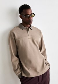 Beige long-sleeve polo shirt with a collar, featuring a subtle logo on the chest, and ribbed cuffs. Worn with dark-colored pants.