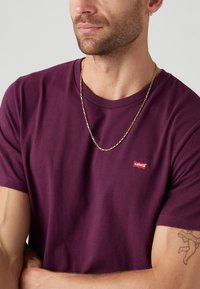 Violet trumpas rankovėmis marškinėliai, pagaminti iš medvilnės, su mažuoju raudonu Levi's logotipu ant priekio ir auksine grandinės apyranke.