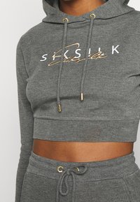 Sudadera recortada gris hecha de una tela suave, con capucha ajustable, texto del logo en dorado y blanco, y acentos en dorado en los cordones.