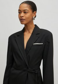 Black blazer with long sleeves, deep V-neck, wrap design, and belted waist. Features a single white-striped breast pocket. Smooth texture.