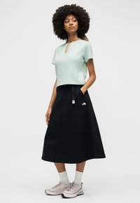 Γυναίκα με αφρο-χτένισμα που φοράει ανοιχτό μπλε κοντομάνικη μπλούζα, μαύρη μέση φούστα με λογότυπο, λευκές κάλτσες και ανοιχτογκρι αθλητικά παπούτσια, στέκεται με το χέρι στην τσέπη.