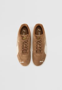 Braune Wildledersneaker mit einem weißen Seitenpaneel, runder Zehenpartie, Schnürdesign und goldenen Akzenten auf der Zunge und der Markenlogo.