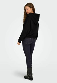 Felpa nera con cappuccio e vestibilità ampia, abbinata a jeans skinny blu scuro. Modella con stivaletti neri, che mostrano una texture naturale nei tessuti.