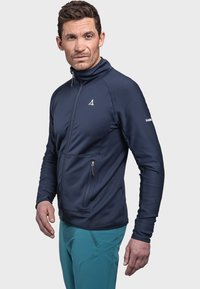 Marineblaues Fleece-Jacke mit Reißverschluss und hohem Kragen, strukturierter Oberfläche, seitlichen Reißverschlusstaschen sowie einem Logo auf der Brust und dem Ärmel. Kombiniert mit türkisfarbenen Hosen.