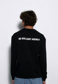 Czarna koszula z długim rękawem z białym napisem na plecach "NO BULLSHIT AGENCY." Wykonana z bawełny, o luźnym kroju.