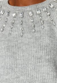 Grauer Strickpullover mit Kristallverzierungen entlang des Ausschnitts. Die Textur ist weich und hat ein durchgängiges Rippmuster im Stoff.