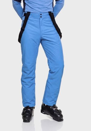Person trägt hellblaue Skihose mit schwarzen Hosenträgern und schwarzen Skischuhen und steht vor einem einfarbigen Hintergrund.