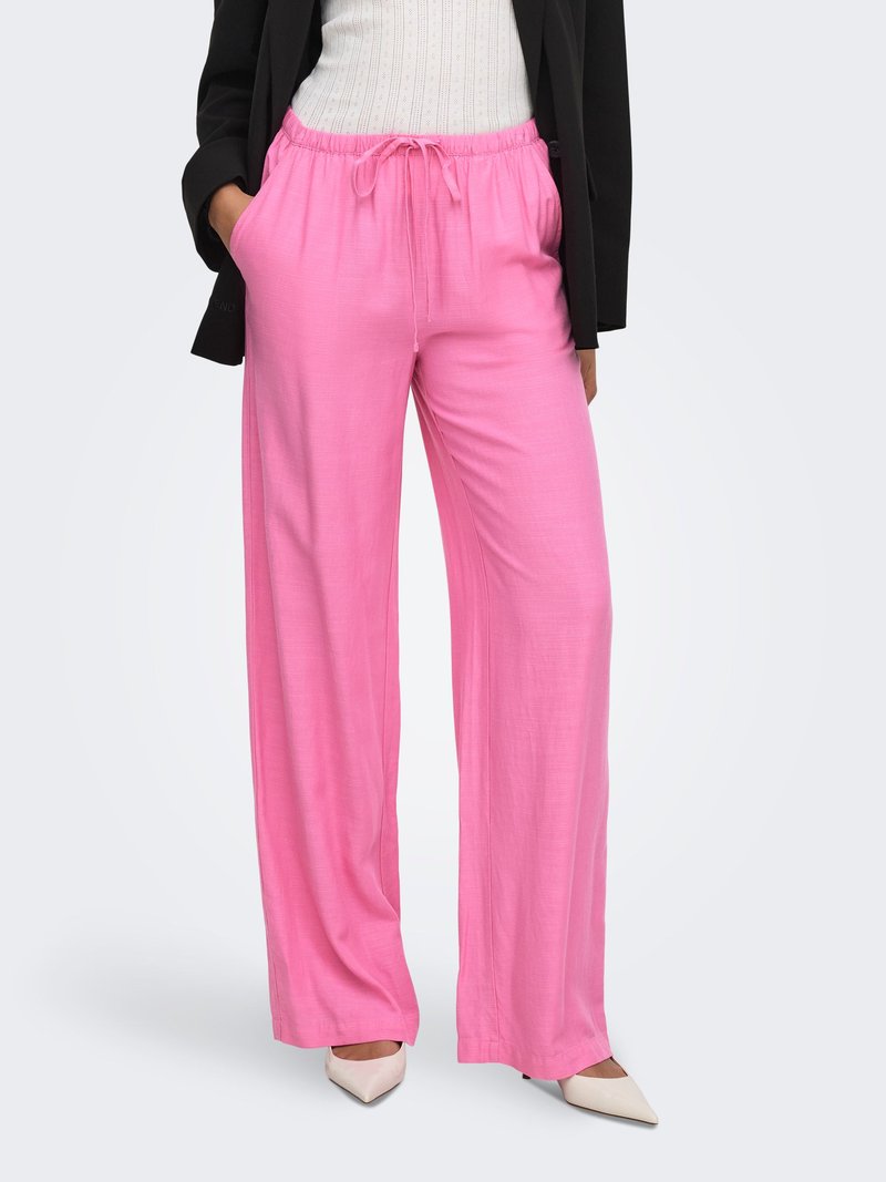 Donna che indossa pantaloni rosa ampi con vita a coulisse, scarpe bianche appuntite, top bianco e blazer nero, in piedi con le mani nelle tasche.