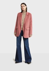 Blazer de mezcla de lana rosa de gran tamaño con solapas de muesca, dos bolsillos frontales, llevado sobre un cuello alto de punto con estampado, combinado con jeans de campana azules.