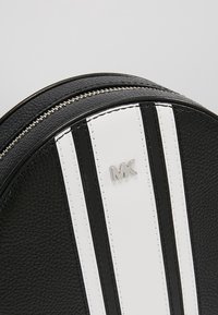 Borsa rotonda nera in pelle con strisce bianche, dettaglio del logo argentato e superficie testurizzata. Presenta una chiusura a zip lungo la parte superiore.