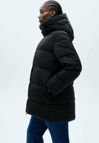 Geacă puf cu quiltare neagră, cu glugă înaltă, buzunare laterale și închidere cu fermoar, purtată peste blugi din denim albastri.