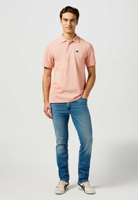 Polo shirt rosa a maniche corte con colletto e un piccolo logo, abbinato a jeans blu chiaro e sneaker crema con dettagli neri.