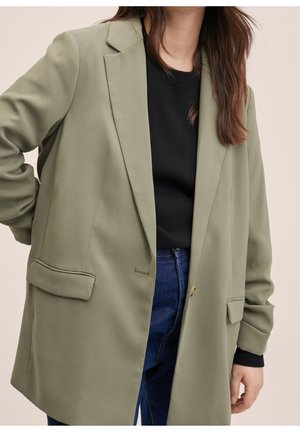 Osoba ubrana w oliwkowozielony blezer na czarnym swetrze i niebieskich dżinsach, stojąca z jedną ręką na biodrze na neutralnym tle.