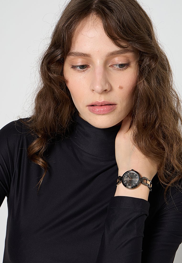 Montre en acier inoxydable noir avec un cadran rond et des accents argentés. Le bracelet présente un design à maillons. Portée sur un poignet avec un col roulé noir.