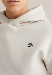 Kreaminvärinen huppari, jossa on pehmeä rakenne ja etupussitasku. Vasemmassa rintakohdassa on musta brodeerattu linnun logo.