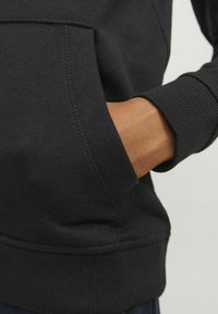 Črna pulover s smooth teksturo, rebrastimi manšeti in kenguru žepom. Ima detajle šivanja in sproščen kroj.