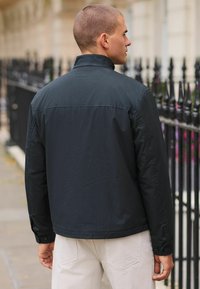Black lightweight jacket with a high collar, buttoned cuffs, and a smooth texture, worn over light-colored pants, viewed from behind.
