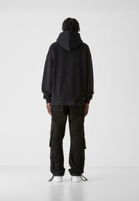 Melna kapuce jaka ar brīvu piegriezumu un ribotu apakšmalu, kombinēta ar melniem biksēm ar lielām sānu kabatām. Izskatu papildina baltas kedas.