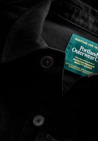 Melna kordurojā tērpa apkakle ar sudraba pogu un zaļu etiķeti, uz kuras ir rakstīts "Portland Outerwear Co." un sniegta informācija par audumu un kvalitāti.