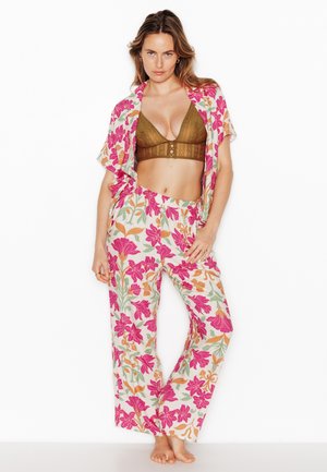 Donna in piedi a piedi nudi che indossa un bralette di pizzo marrone e una camicia aperta e pantaloni coordinati con stampa floreale con fiori rosa, arancioni e verdi su sfondo bianco.