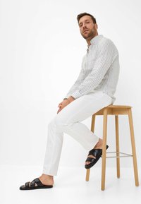 Gestreiftes Button-Down-Hemd in hellen Farben, weiße Slim-Fit-Hose und schwarze Sandalen mit mehreren Riemen. Modell sitzt auf einem Holzstuhl.